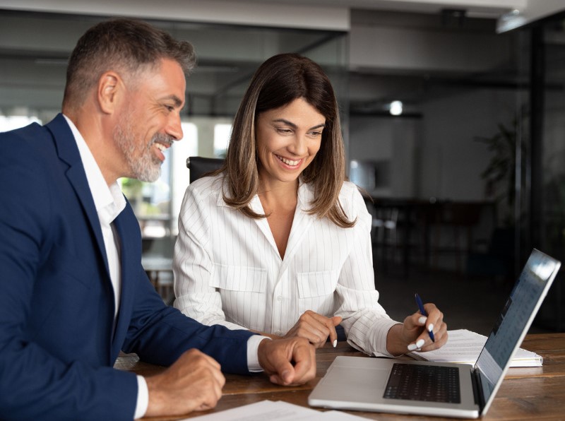 Mature business man mentoring a business woman discussing project on laptop in office around a table.