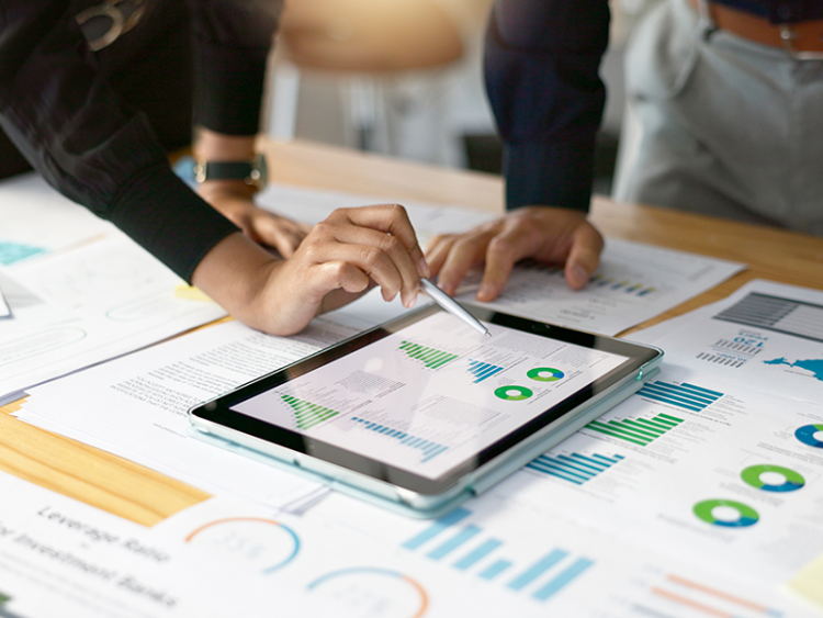 Two people standing over table, we see their hands, one is pointing to graphs on an iPad